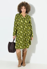Frau mit lockigem Haar, die ein grünes, floral gemustertes Langarmkleid und beigefarbene Wildleder-Stiefel bis zum Knie trägt, während sie eine dunkelbraune, geflochtene Handtasche hält.