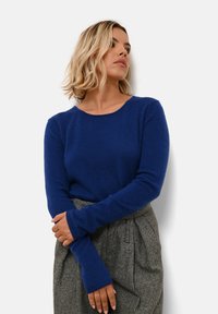 Modrý pletený svetr s dlouhými rukávy, kulatým výstřihem a měkkou texturou. Stylizován s šedými kalhotami s vysokým pasem pro kontrast.