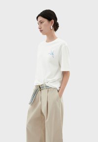 Witte t-shirt met korte mouwen en een blauw grafisch ontwerp op de borst, gecombineerd met beige high-waisted, wijde broek en een geblokte riem.