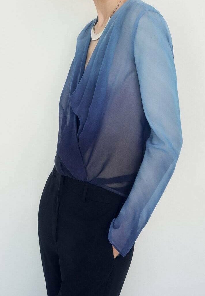 Translucent blouse featuring a gradient from light blue to navy, with a draped front, long sleeves, and a relaxed fit, paired with black pants.
