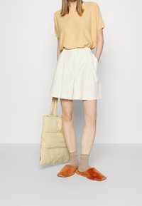 Jasnożółta bluzka z krótkim rękawem, białe plisowane szorty, beżowa puchowa torba na ramię, brązowe skarpetki z falbanką oraz pomarańczowe, puszyste buty wsuwane.