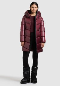 Burgundijska puffer jakna s kapuljačom, koja ima kvadratni dizajn i sjajni završetak. Crne snježne čizme s teksturiranim izgledom i zatvaranjem na šnuru.