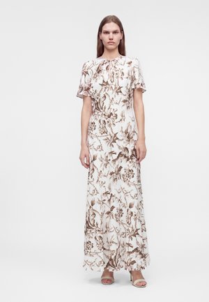 Femme debout portant une longue robe blanche avec des motifs floraux marron et des manches courtes, avec des talons ouverts beiges.