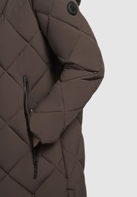 Brun quiltet pufferjakke med en lynlås lomme og et sort logo patch. Stoffet ser blødt ud med en glat tekstur.