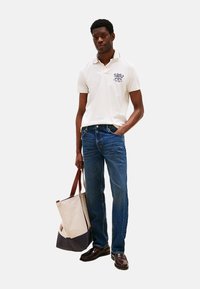 Camiseta polo blanca con un logo bordado azul, combinada con jeans azules. El modelo sostiene una bolsa de tote beige y navy y lleva zapatos oscuros.