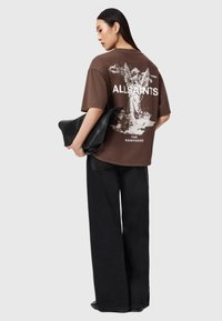 Camiseta gráfica marrón con estampado de ángel, diseño oversize, mangas cortas, combinada con pantalones negros de pierna ancha, sosteniendo un bolso de mano negro.