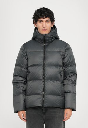 Graue Daunenjacke mit Kapuze, die über ein gestepptes Design, einen Frontreißverschluss und eine leicht glänzende Textur verfügt. Getragen über dunkelgrauen Hosen.