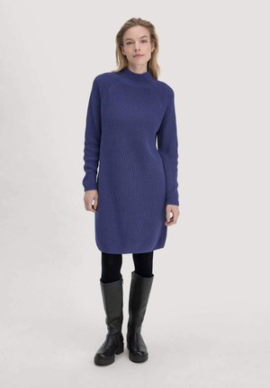 Vrouw in een lang mouwen blauwe gebreide jurk, zwarte leggings en zwarte kniehoge laarzen, staande tegen een effen lichte achtergrond.