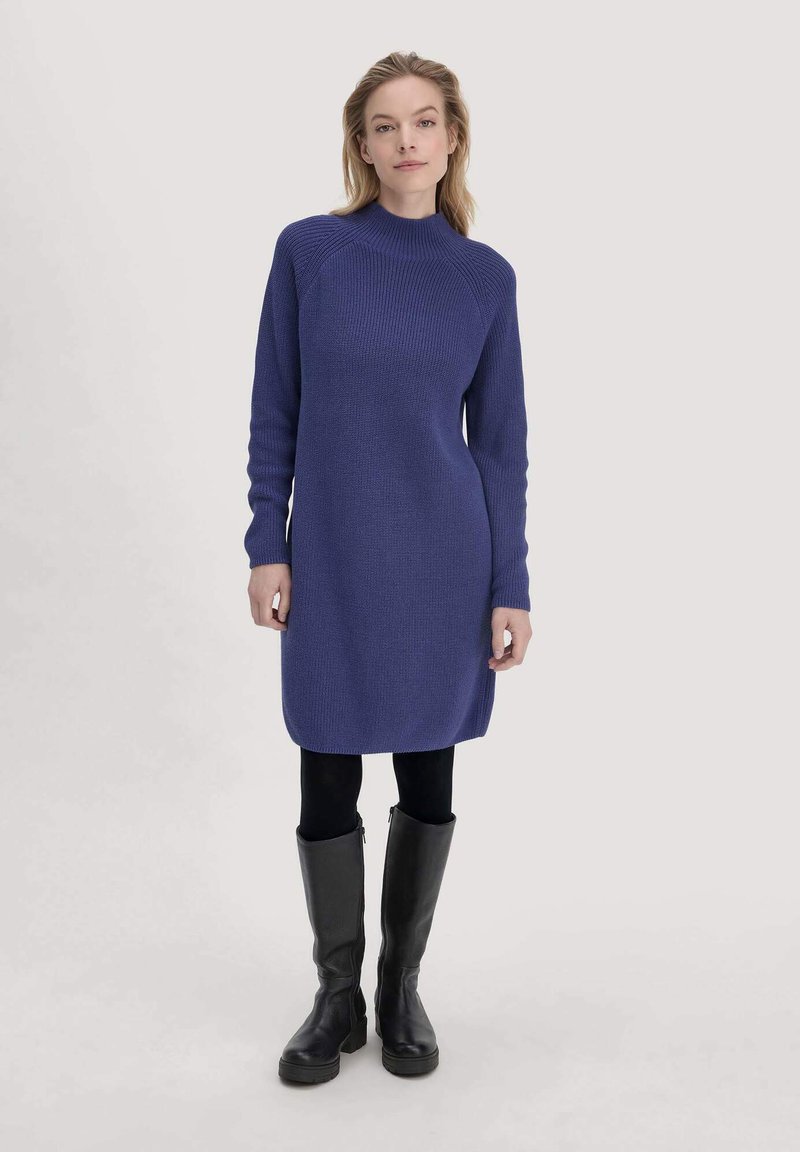 Donna che indossa un vestito di maglia blu a maniche lunghe, leggings neri e stivali neri fino al ginocchio, in piedi davanti a uno sfondo chiaro e semplice.