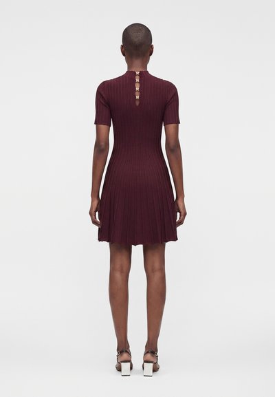 Personne portant une robe à manches courtes, côtelée, de couleur rouge foncé avec des détails de boutons dorés dans le haut du dos, associée à des sandales blanches à talons blocs.
