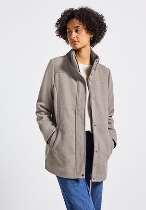 Manteau en mélange de laine gris avec un col haut, des boutons pression, des poches latérales et une finition texturée. Le modèle le porte par-dessus un haut clair et un jean foncé.