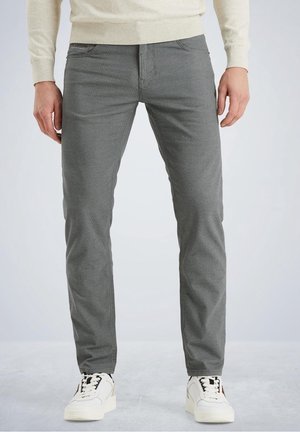 Mężczyzna ubrany w szare, dopasowane spodnie o fakturze oraz białe trampki, stojący na tle jednolitego, jasnego tła.