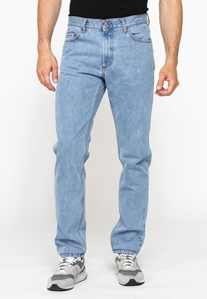 Uomo che indossa jeans azzurri a gamba dritta, camicia nera e sneaker grigie bianche, in piedi davanti a uno sfondo bianco uniforme.