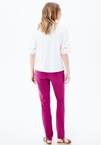 Top bianco a maniche corte con ritagli ondulati sui polsini, abbinato a pantaloni fucsia aderenti. Il modello si trova su uno sfondo neutro.