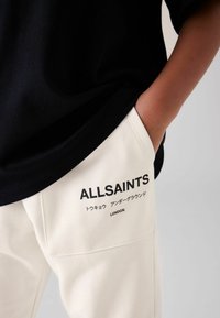 Sort bomulds T-shirt parret med cremefarvede bukser med et logo og tekstprint. Blødt stof med en afslappet pasform, sidelommer.