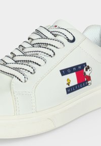Baskets blanches avec lacets à rayures noires, logo Tommy Hilfiger et personnages de dessin animé Snoopy et Woodstock sur le côté.