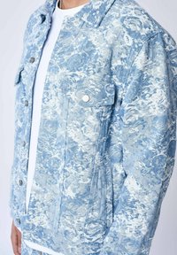 Giacca di jeans azzurro chiaro con un motivo astratto testurizzato, finiture in argento, colletto a punta e tasche applicate frontali.