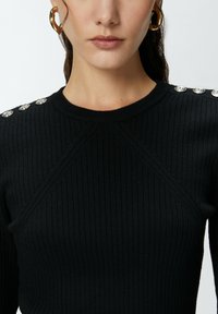 Maglione nero a coste con maniche lunghe e scollo rotondo, con bottoni metallici decorativi sulle spalle e dettagli testurizzati.