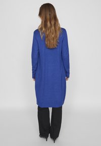 Cárdigan de punto largo y azul con cuello redondo, detalles acanalados y un corte relajado. Lleva puesto sobre pantalones negros, mostrando un color sólido y una textura suave.