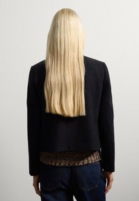 Veste en mélange de laine noire avec épaules structurées, ourlet court et pull en maille texturé marron en dessous, associé à un jean en denim bleu.