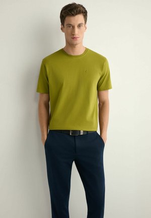 Młody mężczyzna z krótkimi brązowymi włosami, ubrany w oliwkowo zieloną koszulkę z krótkim rękawem i ciemnoniebieskie spodnie, stoi z rękami w kieszeniach na tle białej ściany.