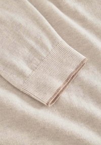 Beiger Strickpullover in Nahaufnahme, mit gerippten Manschetten und einem hellbraunen Akzent, der eine weiche Textur und einen glatten, leichten Stoff zeigt.