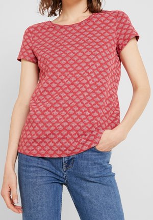 Kvinde iført en kortærmet rød T-shirt med mønster og blå jeans med den ene hånd i lommen mod en ensfarvet baggrund.