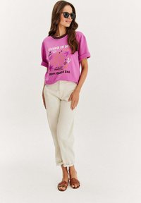 Růžové grafické tričko s ohrnutými rukávy, zdobené barevným potiskem. V kombinaci se smetanovými zužujícími se kalhotami a hnědými sandály na řemínky.
