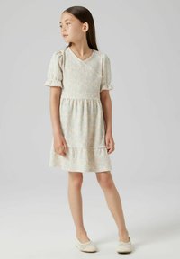 Leicht strukturiertes Kleid in sanften Farben, mit einem V-Ausschnitt, kurzen Puffärmeln, einer gerafften Taille und einem gestuften Saum. Kombiniert mit hellen Schuhen.