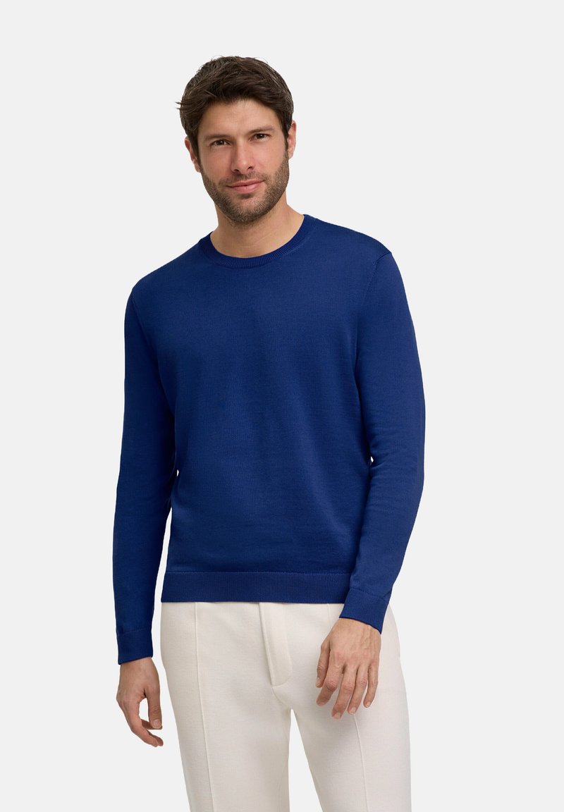 Blauer Strickpullover mit rundem Halsausschnitt, langen Ärmeln, strukturiertem Stoff und gerippten Bündchen sowie Saum. Getragen mit hellen Hosen.