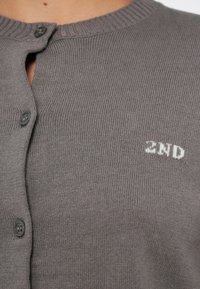 Szürke kötött kardigán kerek nyakkivágással, gombos lezárással és egy kis fehér "2ND" logóval a bal oldalon. Puha textúra bordázott mintával.