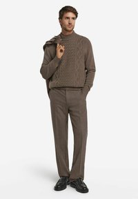 Brązowy sweter z dzianiny o fakturowanym wzorze w warkocze, z prążkowanym dekoltem, zestawiony z jasnobrązowymi spodniami i czarnymi butami. Dodatki obejmują sweter zarzucony na ramię.