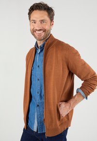 Cárdigan marrón con cremallera frontal, hecho de material suave, que presenta puños acanalados y un ajuste casual sobre una camisa de mezclilla azul con botones.