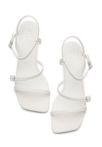 Sandales blanches à lanières, avec un bout carré, en matériau lisse. Elles présentent de fines lanières à la cheville et aux orteils, une boucle réglable et un talon plat.