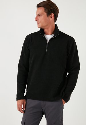Pullover in pile nero con zip parziale, caratterizzato da una superficie texture e colletto alto, abbinato a una t-shirt bianca sottostante.