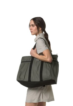 Kobieta o brązowych włosach związanych w kucyk, niosąca dużą zielono-czarną torbę K-Way na ramieniu, ubrana w beżową sukienkę z krótkim rękawem.