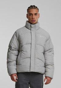 Chaqueta acolchada gris claro con alto cuello, diseño acolchado y logotipo sutil. Presenta un acolchado grueso y cierre con cremallera en la parte delantera.