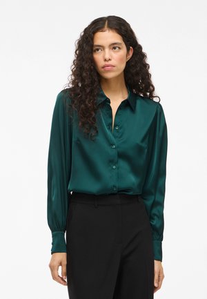 Teal satiinist blouse, millel on krae, nööbiga ees ja pikad varrukad, silmapaistva sileda tekstuuri ja ühe rinna taskuga.