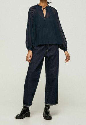 Frau trägt eine plissierte, transparente dunkelblaue Bluse mit Schleifenkragen, dunkle hoch taillierte weite Jeans und schwarze klobige Knöchelstiefel.
