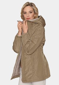 Veste légère beige avec capuche, fermeture éclair à l'avant et poignets élastiques. Texture lisse avec doublure intérieure et poches latérales.