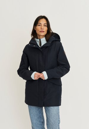 Vrouw met een donkerblauw winterjack met capuchon over een lichtblauwe trui en lichtblauwe jeans, staand met samengevouwen handen.