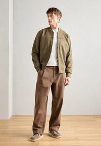 Olive green bomber jacket with a zip front, cream sweater, brown wide-leg trousers, and beige slip-on shoes, set against a neutral background.
