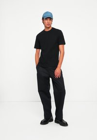 Junger Mann, der eine hellblaue Baseballmütze, ein schwarzes T-Shirt, schwarze Hosen und schwarze Schuhe trägt und mit teilweise in den Taschen stehend.