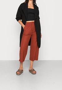 Femme portant un pantalon court couleur rouille, un top tube noir, un gilet ouvert noir et des sandales à enfiler imprimées léopard, se tenant avec une main dans la poche.