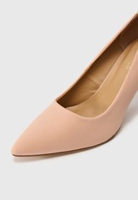 Escarpin à talon haut de couleur rose nude avec un design à bout pointu, dessus en tissu lisse et une texture mate subtile. Design ouvert sur le côté révélant la doublure intérieure.