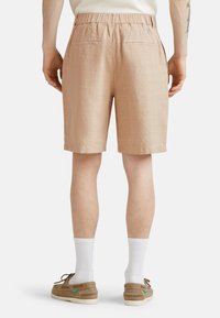 Bermuda color beige chiaro con vita elastica, motivo a quadretti, due tasche posteriori e una vestibilità rilassata, abbinate a scarpe marroni e calzini bianchi.