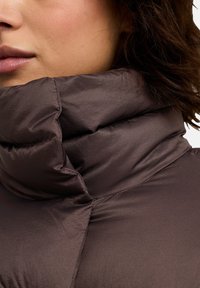 Veste rembourrée marron avec un col haut, présentant une texture lisse et brillante ainsi que des coutures bien marquées, soulignant la chaleur et l'isolation.