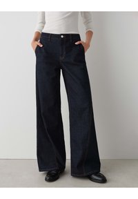 High-waisted, wide-leg jeans in dark denim, featuring contrast stitching, side pockets, and a front button closure. Smooth texture.