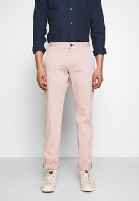 Różowe bawełniane spodnie chino o prostym kroju, z bocznymi kieszeniami i podwiniętymi mankietami, zestawione z granatową koszulą na guziki i białymi trampkami.