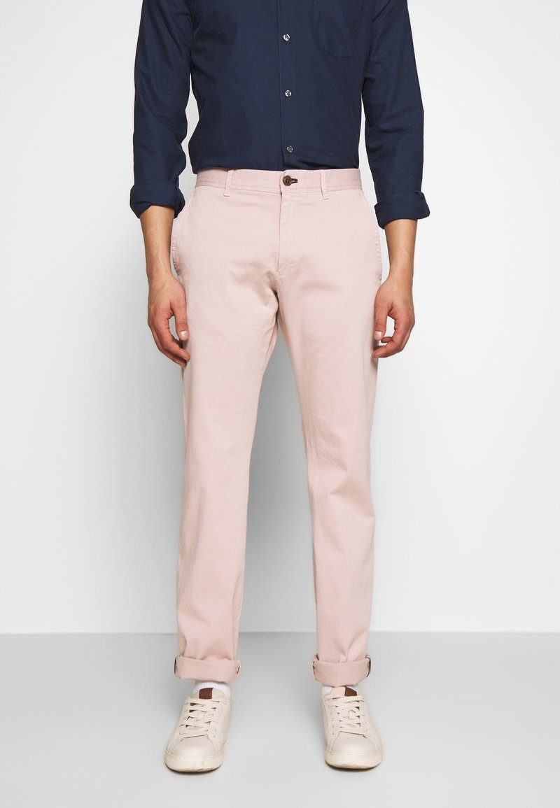 Różowe bawełniane spodnie chino o prostym kroju, z bocznymi kieszeniami i podwiniętymi mankietami, zestawione z granatową koszulą na guziki i białymi trampkami.